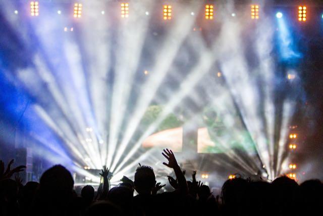 The feature image shows a crowd in front of a stage with bands of lights shooting upward on the stage where a silhouetted person stands